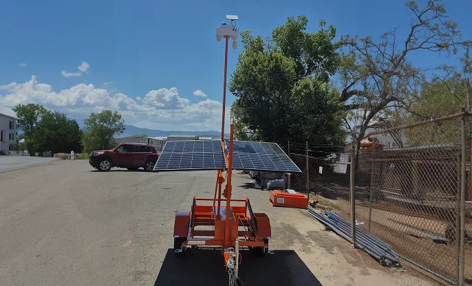 Mobile solar panel trailer with security camera installed, parked on a paved area near a chain-link fence and trees.