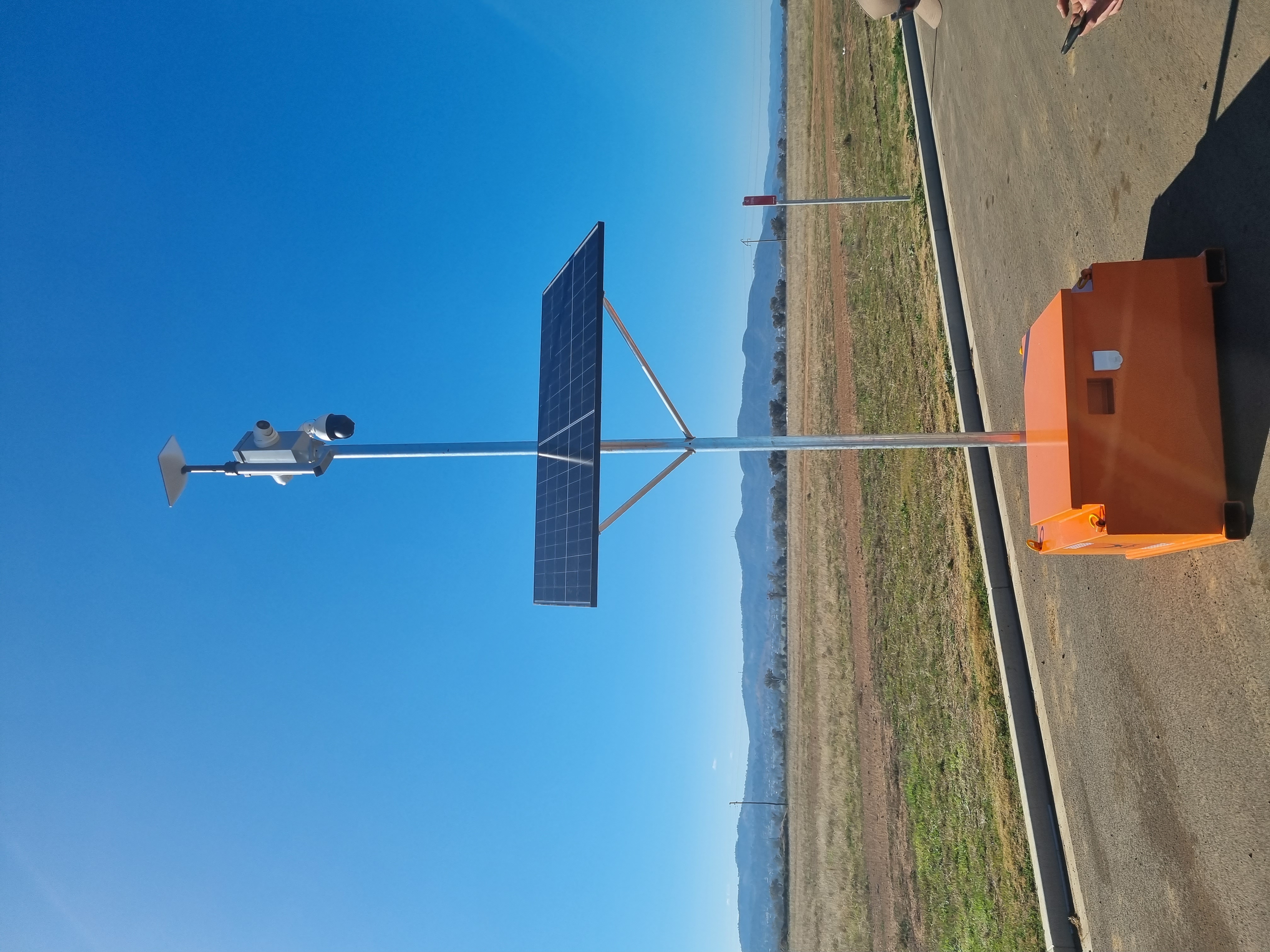Siteguard solar camera unit with a Starlink dish, dome camera and PTZ camera on a mast above an orange steel base, beside open paddocks