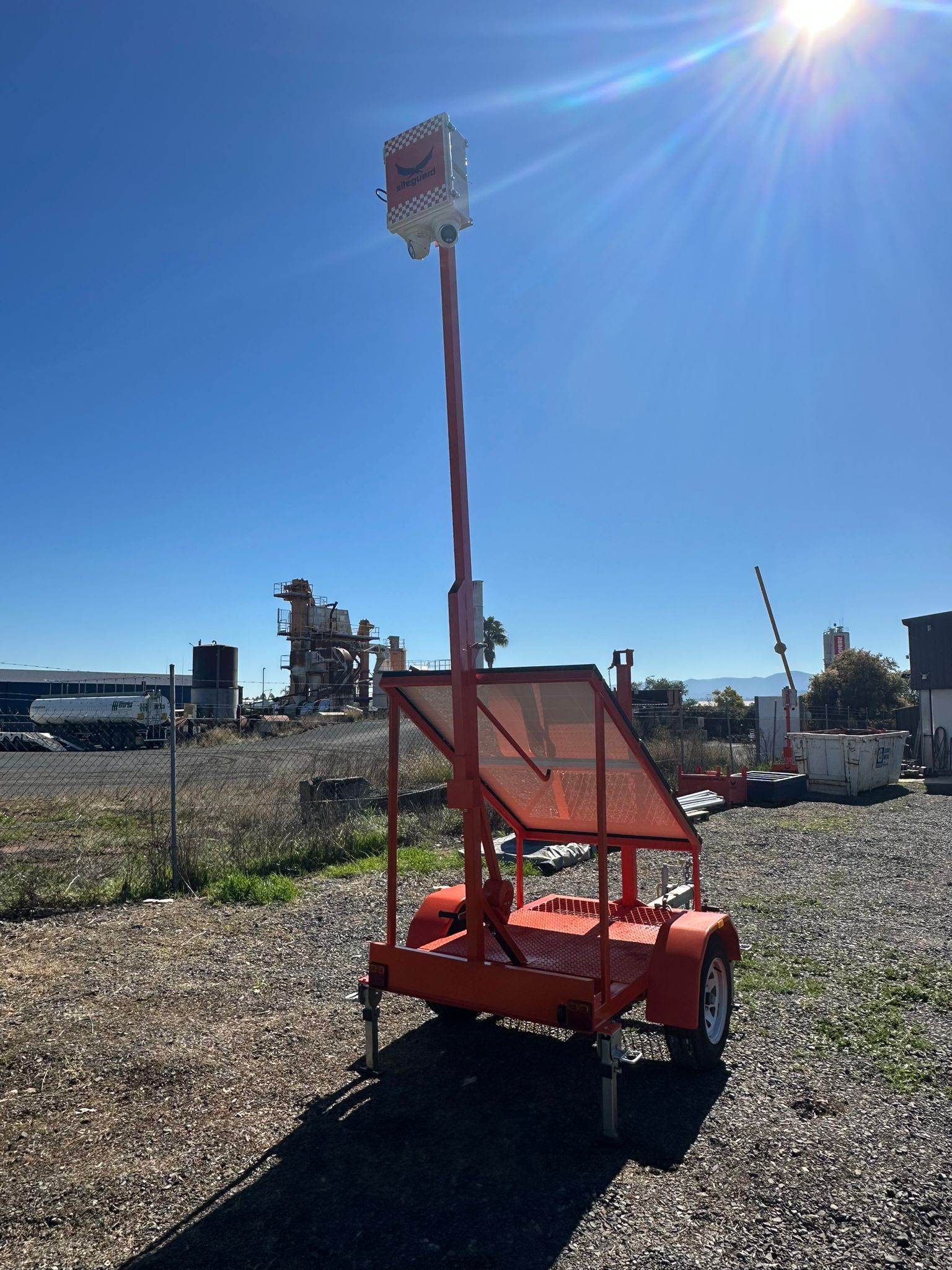 Orange Siteguard solar surveillance trailer with its mast raised and solar panel tilted towards the sun, parked on gravel at an industrial site
