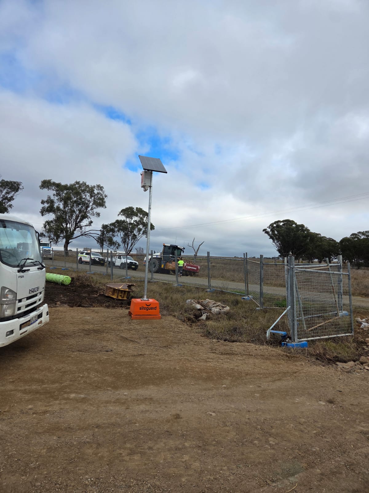 Siteguard solar camera unit on an orange base beside temporary fencing and a site gate, with a truck, utes and a roller on a rural work site