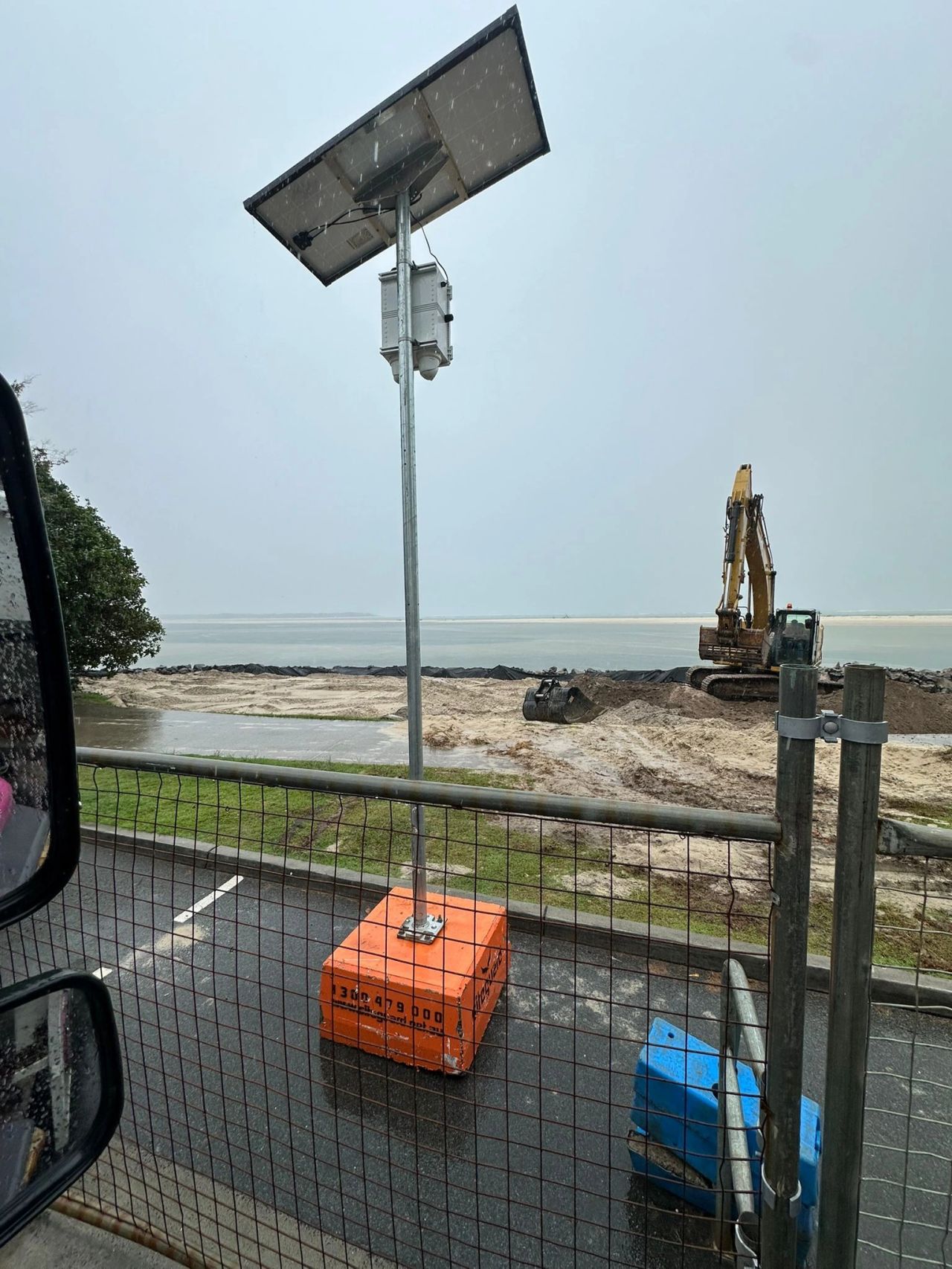 Siteguard solar camera on an orange freestanding base behind temporary fencing, with an excavator working on a sandy foreshore in the rain