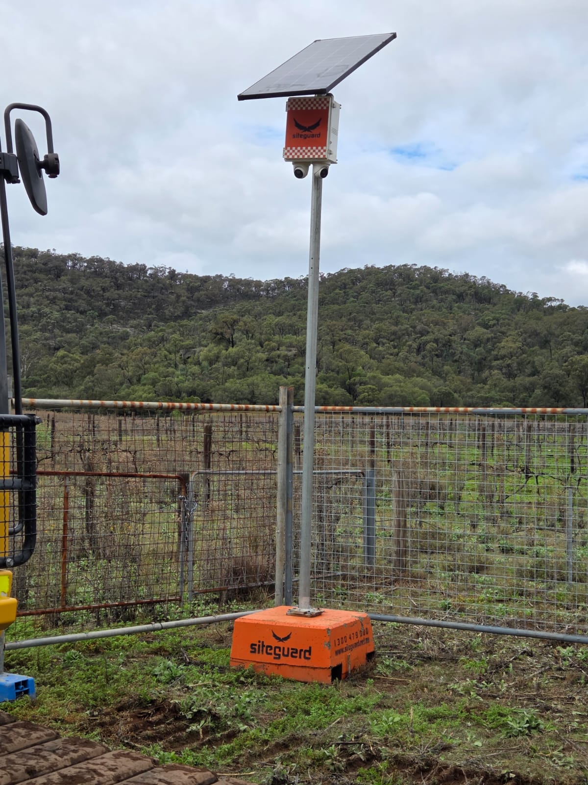 Siteguard solar camera unit with two cameras on a pole and an orange freestanding base, inside a wire boundary fence with wooded hills behind