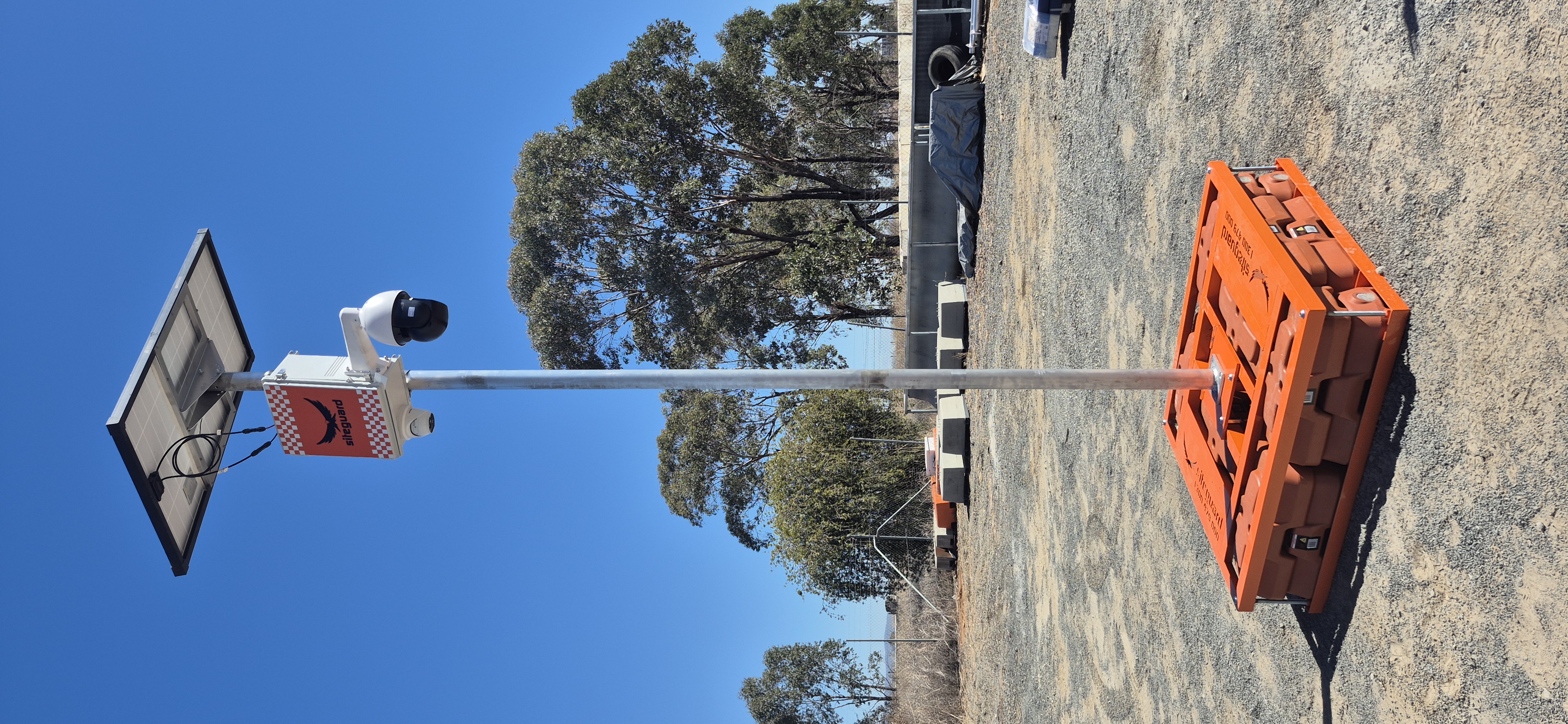 Siteguard solar camera pole with a pan-tilt-zoom dome camera and solar panel on an orange freestanding base in a gravel yard