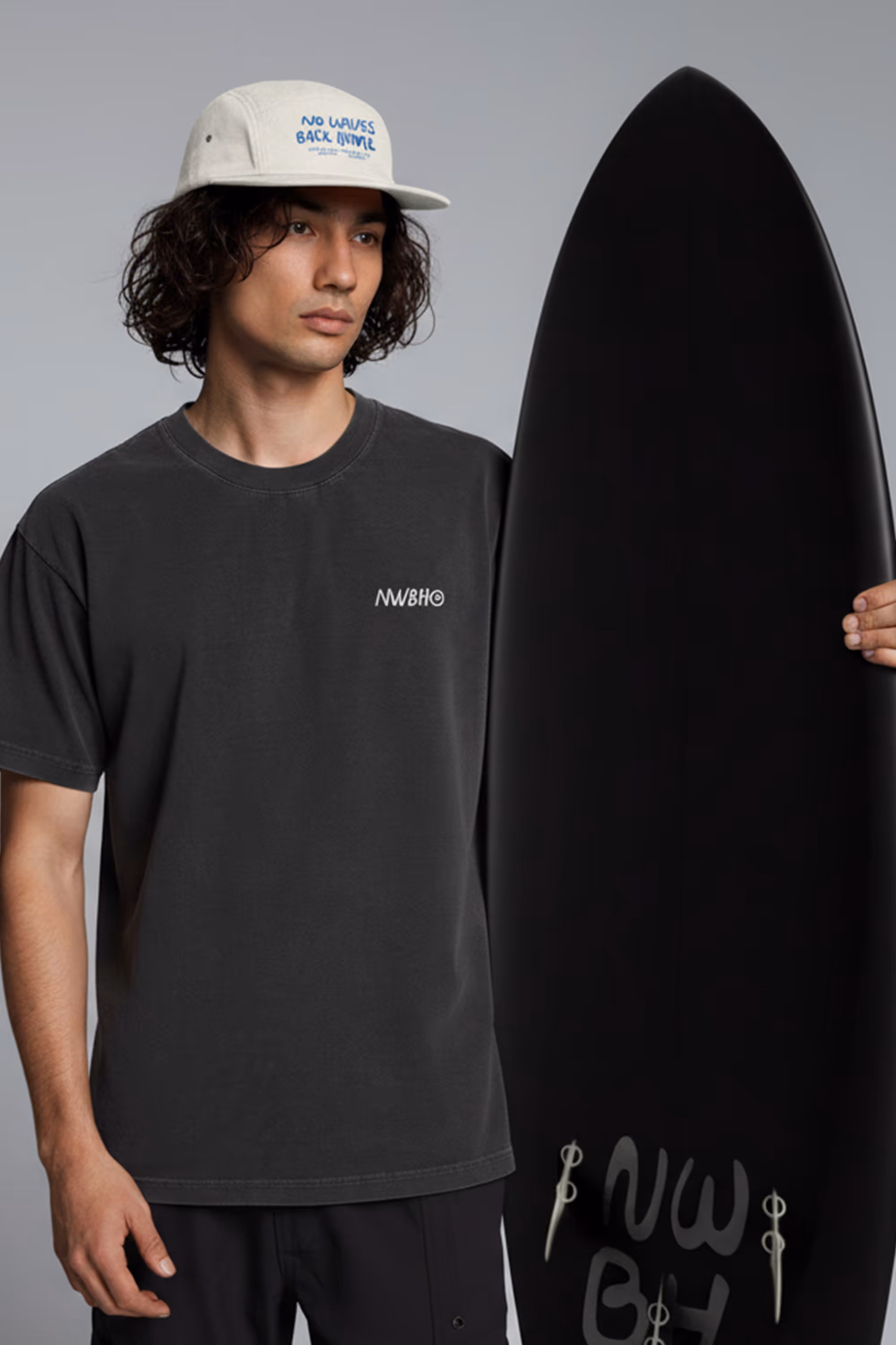 Young man wearing a beige cap and black t-shirt holding a black surfboard with white logo.