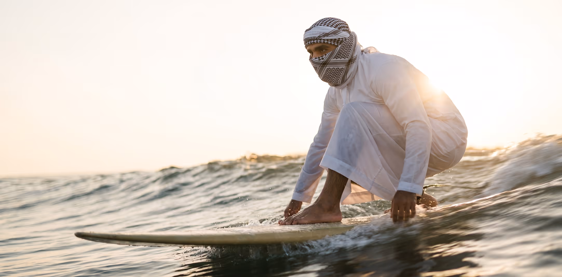 Shirtless young man carrying a black surfboard walking on a sandy beach near the ocean at sunset.