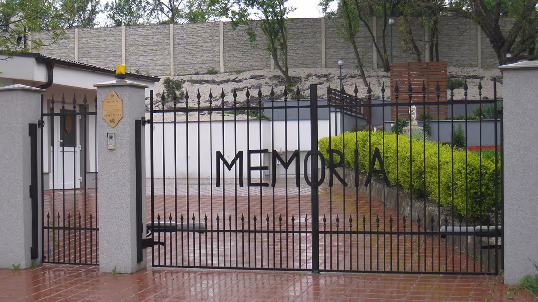 Čierna kovová brána s nápisom MEMORIA pred budovou a zelenými kríkmi.