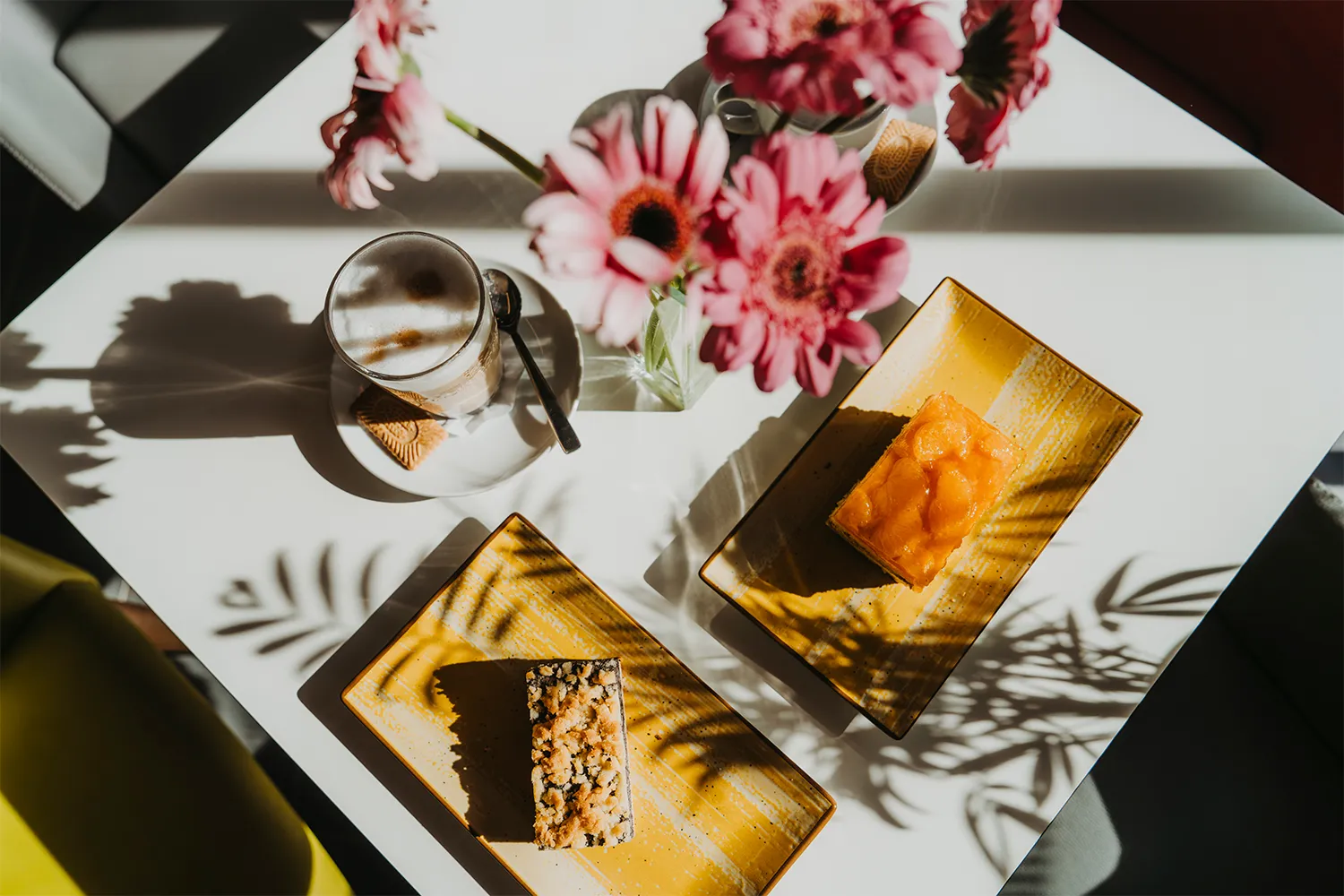 Draufsicht auf einen Tisch mit Cappuccino, frischem Kuchen und sommerlichen Blumen im sonnigen Kurhaus Café Bad Bevensen.