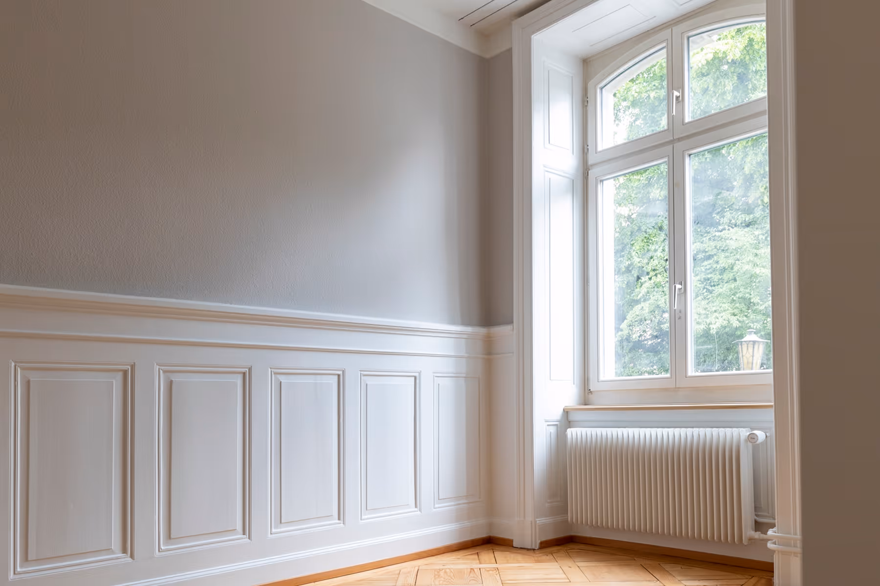 Leerer Raum mit dekorativen Wandpaneelen, grossem Fenster und Heizkörper darunter.