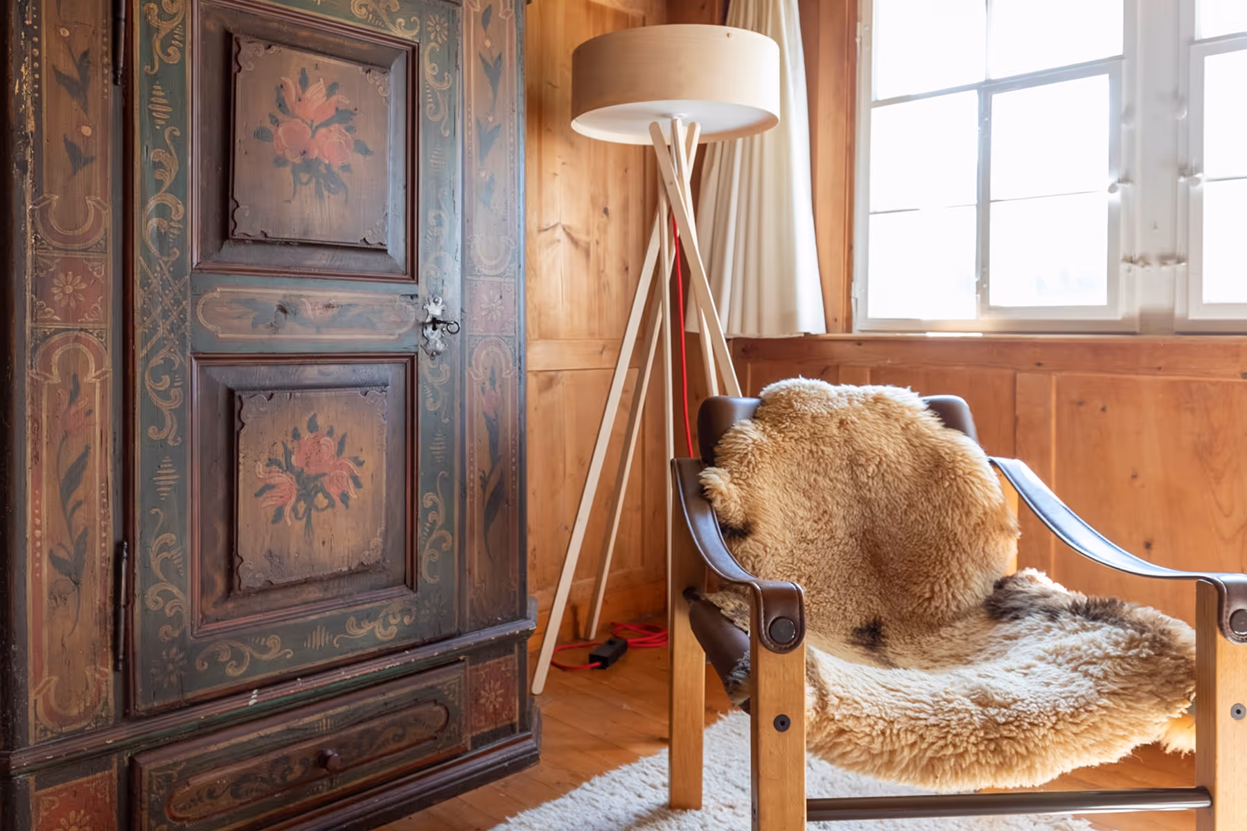 Holzstuhldetail mit braunem Lammfell vor einem Fenster und gemaltem Holzschrank in warmem Zimmer
