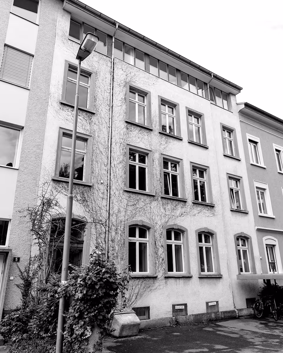 Schwarz-weisses Bild einer mehrstöckigen Wohnhausfassade mit einer Strassenlaterne und Pflanzen im Vordergrund.