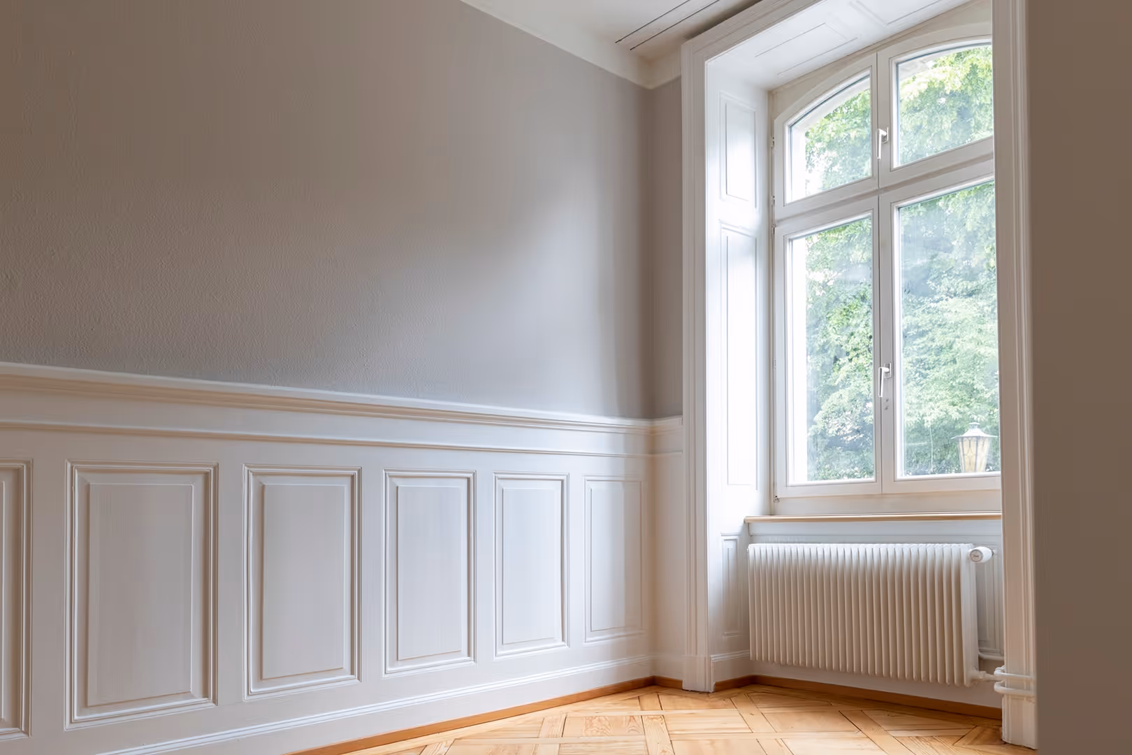 Eleganter leerer Raum mit Holzparkett, weißen Wandverkleidungen und großem Fenster mit Heizung.