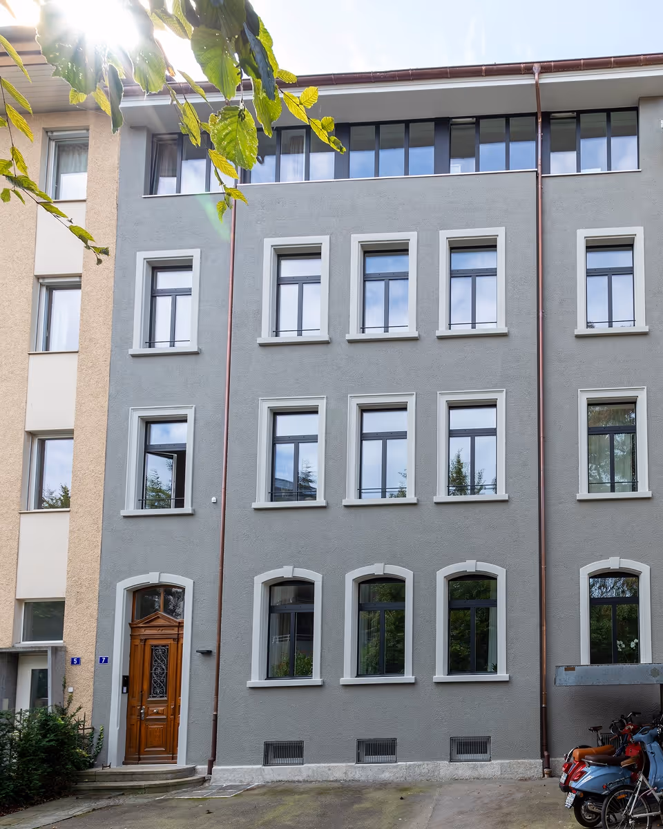 Mehrstöckiges Wohngebäude mit grauer Fassade, brauner Holztür und mehreren Fenstern, Sonnenlicht durch Blätter.