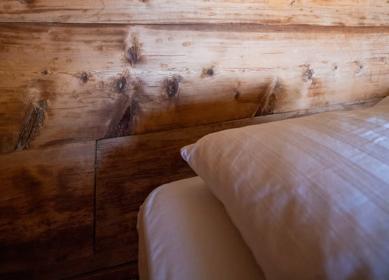 Weisses Kissen auf Bett vor einer rustikalen Holzwand mit sichtbaren Maserungen und Knoten.