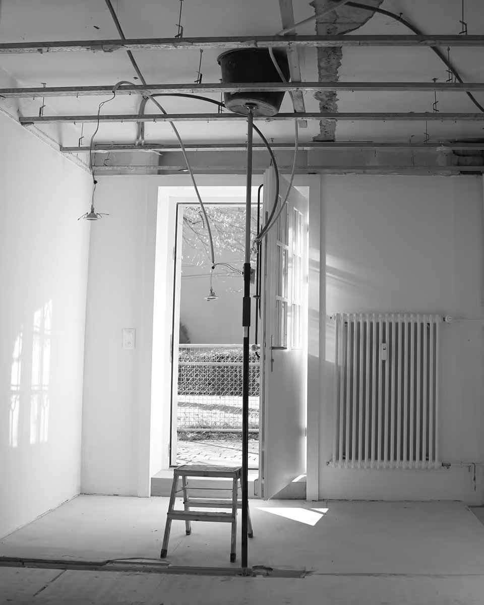 Leerstehender Raum mit offener Tür, Heizkörper an der Wand und Baugerüst mit Kabeln an der Decke.