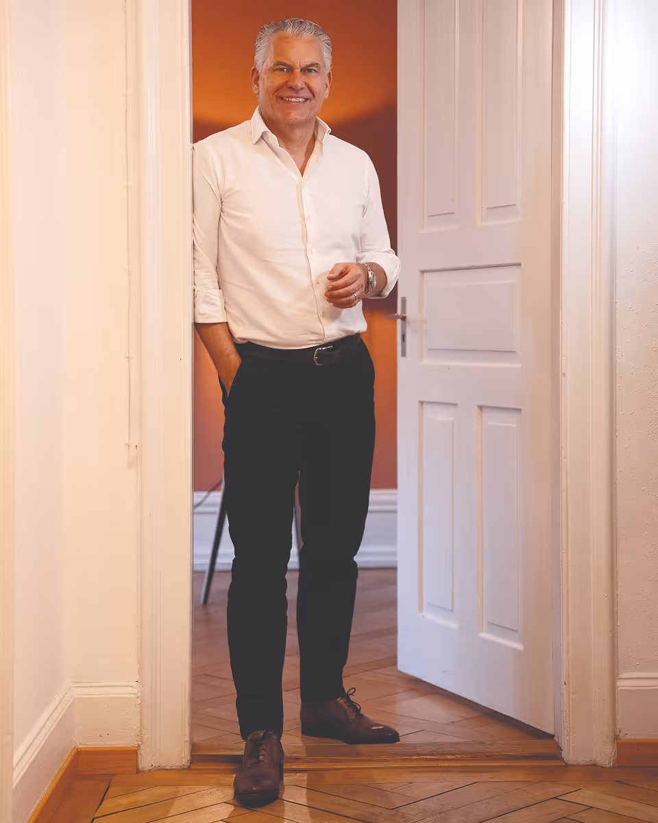 Dr. Philip Meier in weissem Hemd und schwarzer Hose lächelt im Türrahmen eines Zimmers.