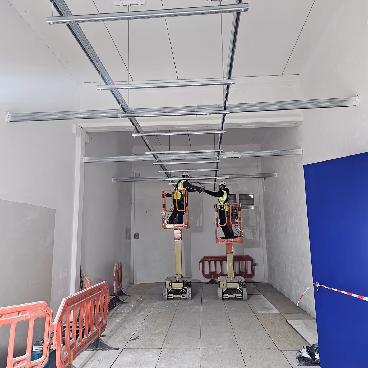 Two construction workers in safety gear on lifts fist bumping inside a white, industrial space.