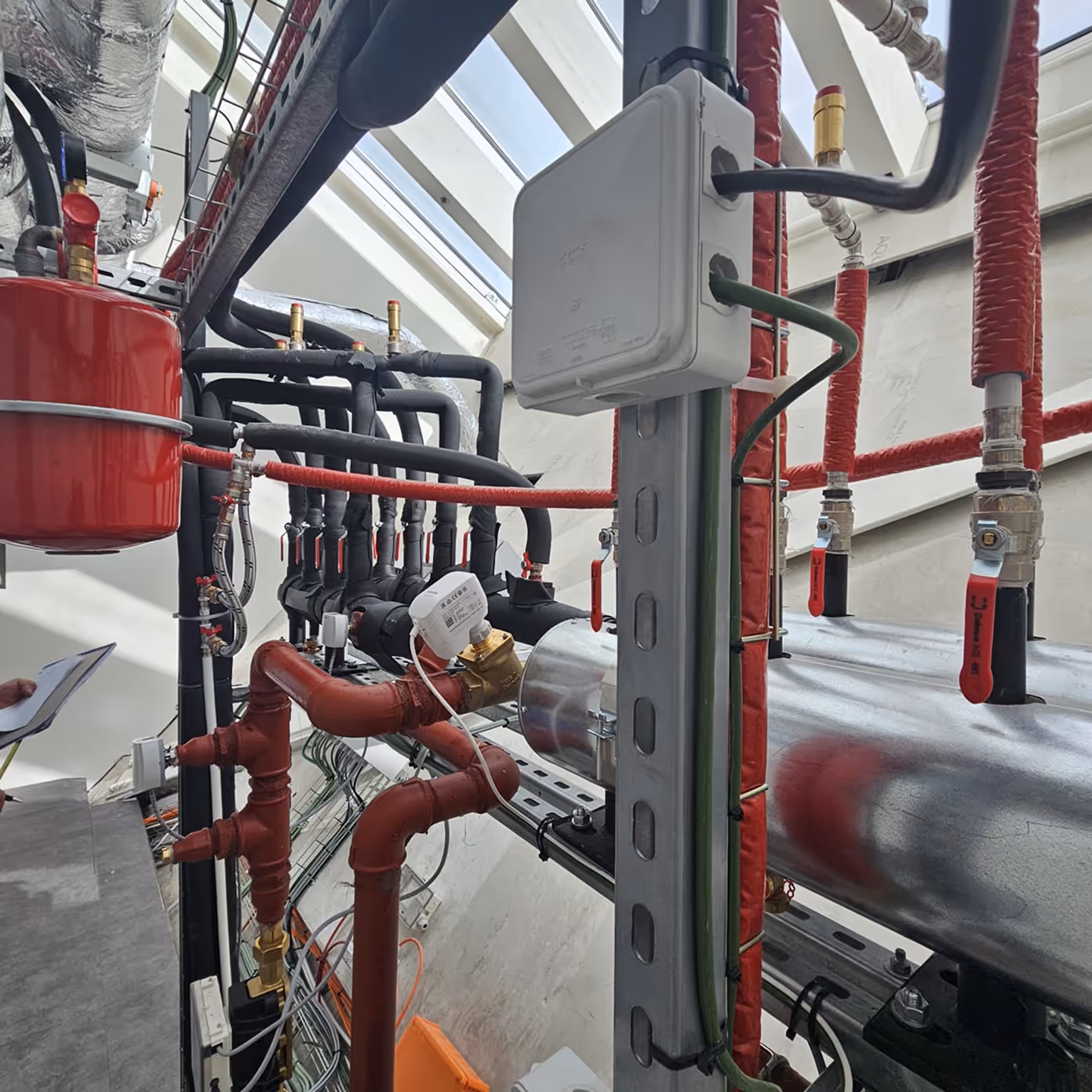Complex industrial piping system with red and black insulated pipes, valves, gauges, and a white control box.