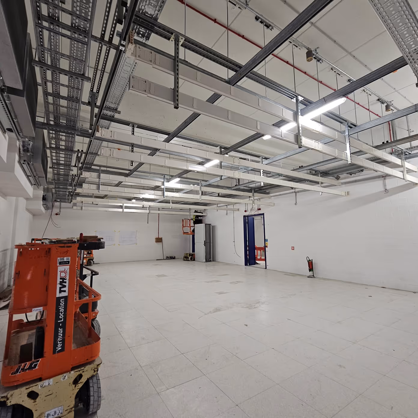 Empty industrial room with white tiled floor, overhead cable trays, and orange construction lift.