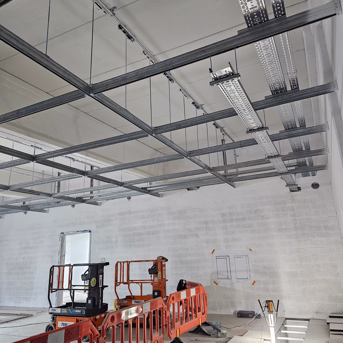 Interior of construction site with suspended metal ceiling frames, lift equipment, and safety barriers.