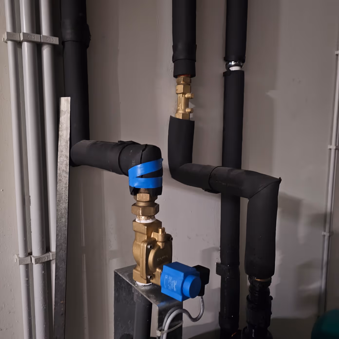 Brass valve with blue actuator connected to black insulated pipes against a gray wall.