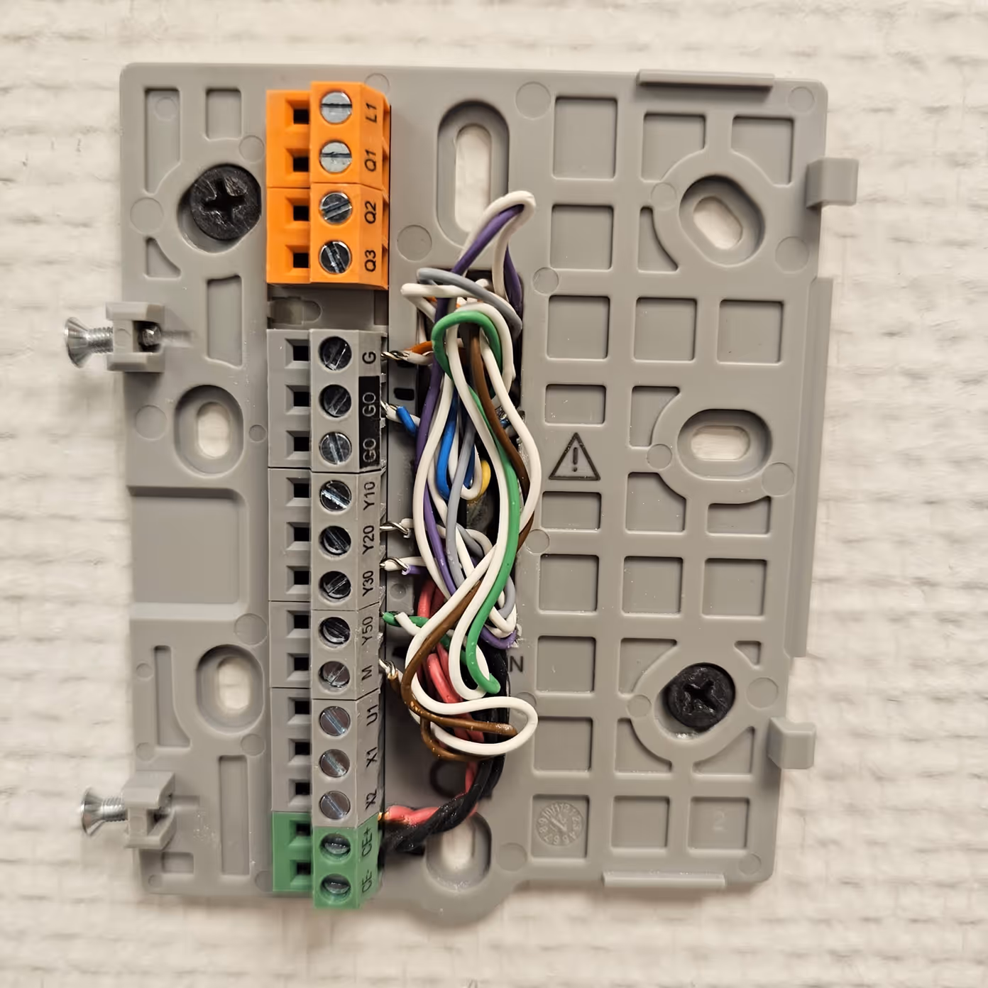 Thermostat wiring panel with multicolored wires inserted into labeled terminals on a gray plastic base.