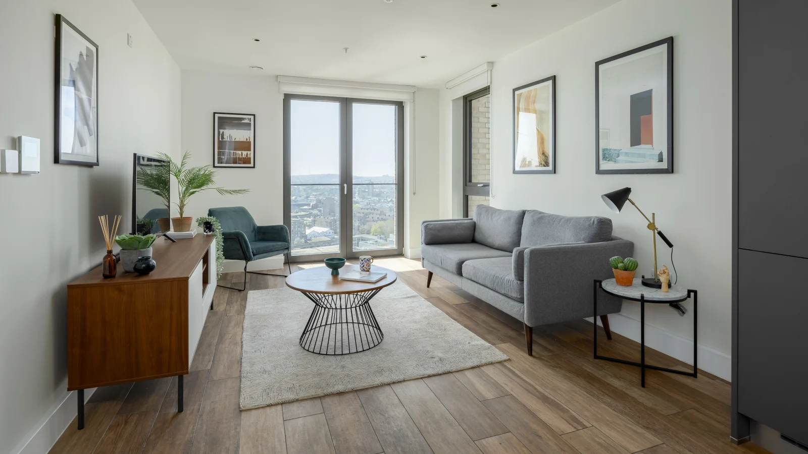 Modern living room with wooden floor, gray sofa, circular coffee table on a light rug, teal armchair, wooden TV stand with plants, floor lamp, and large windows overlooking a cityscape.