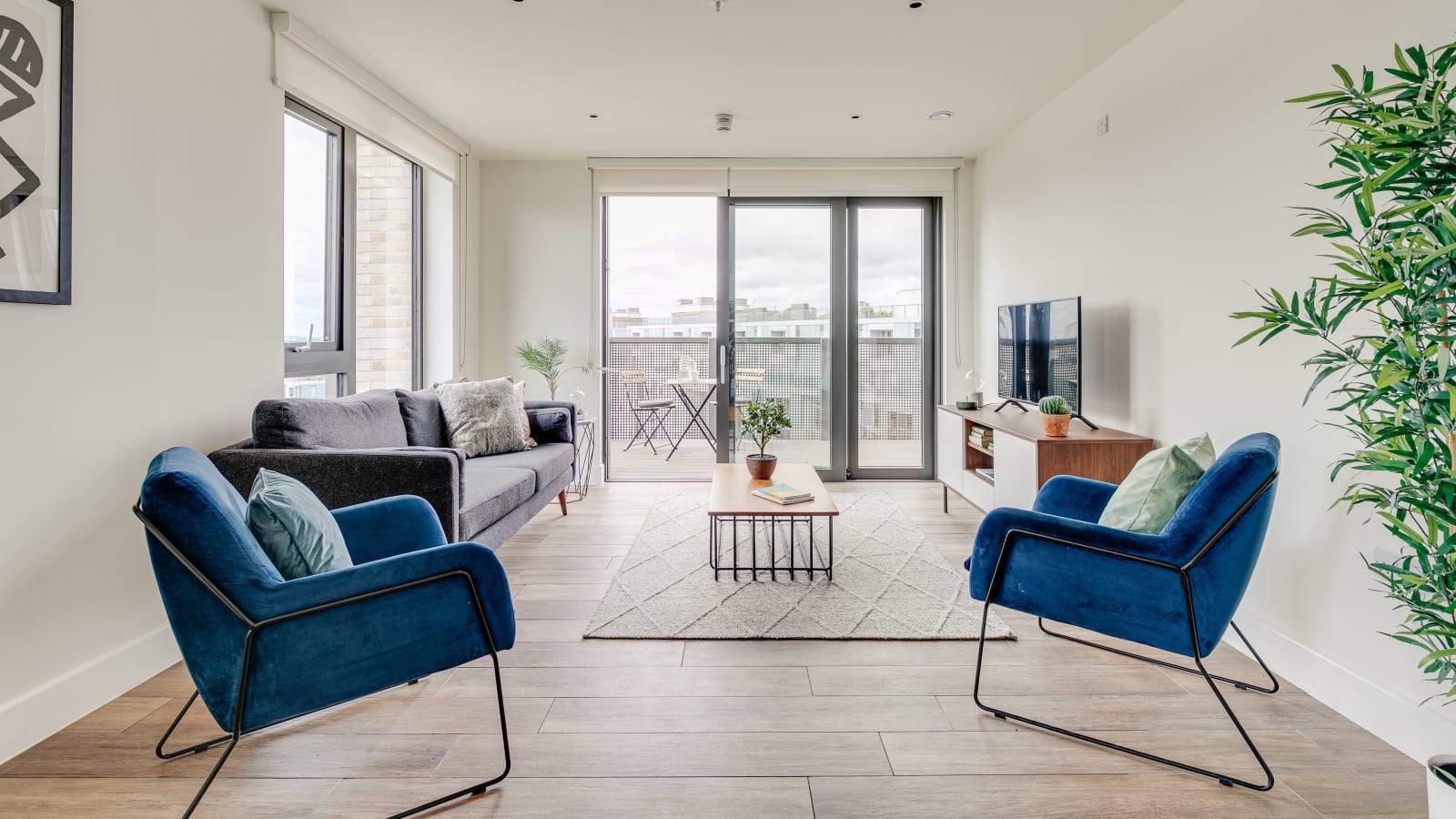 Modern living room with two blue armchairs, a gray sofa, wooden coffee table on a patterned rug, TV on a wooden console, and glass doors leading to a balcony with outdoor seating.