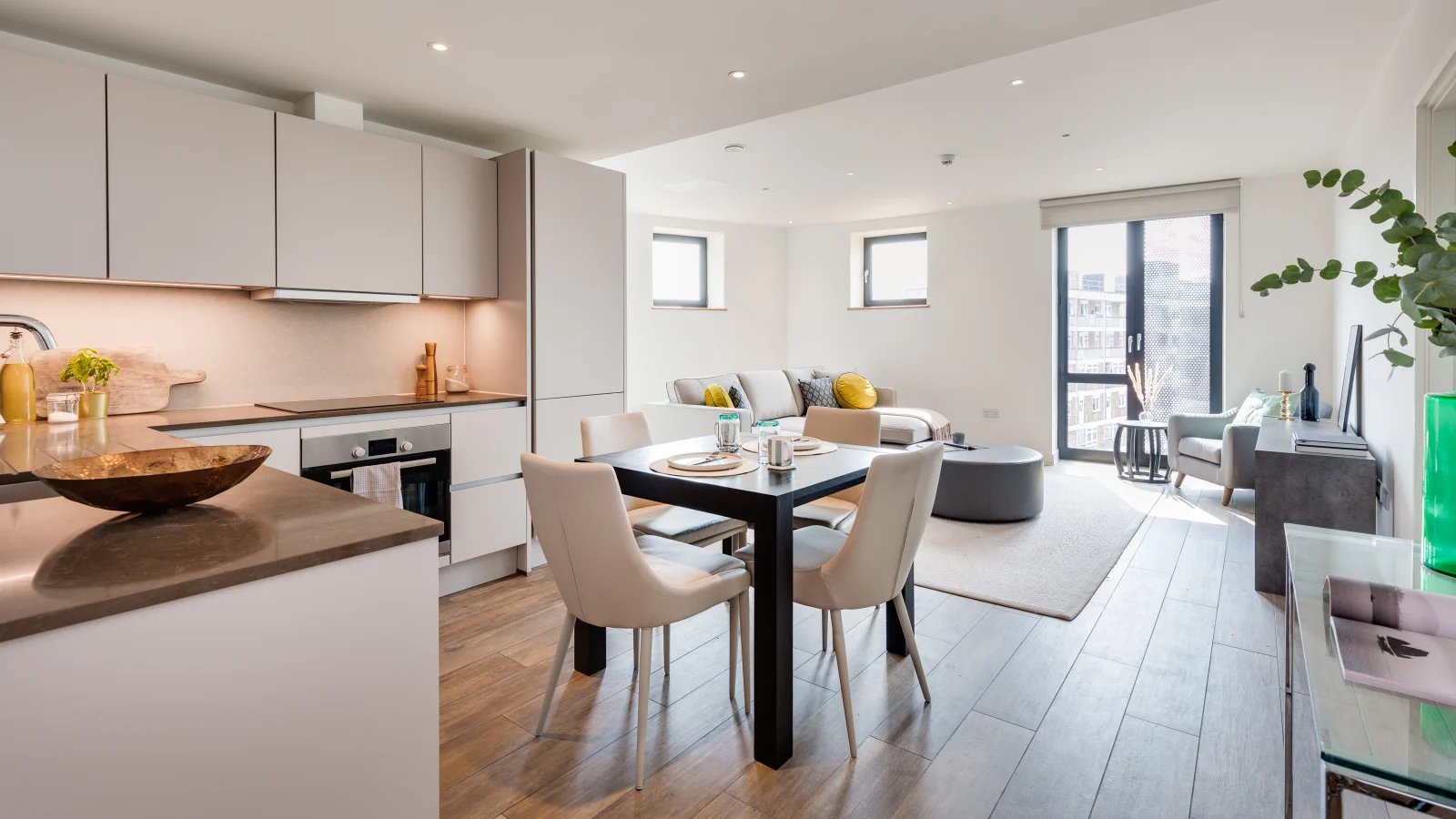 Modern open-plan living space with a sleek kitchen featuring light-colored cabinets and countertops, a black dining table set with four beige chairs and tableware, and a bright seating area with a gray sectional sofa adorned with yellow and patterned cushions, a gray armchair by a large window, and a round ottoman on a light rug.