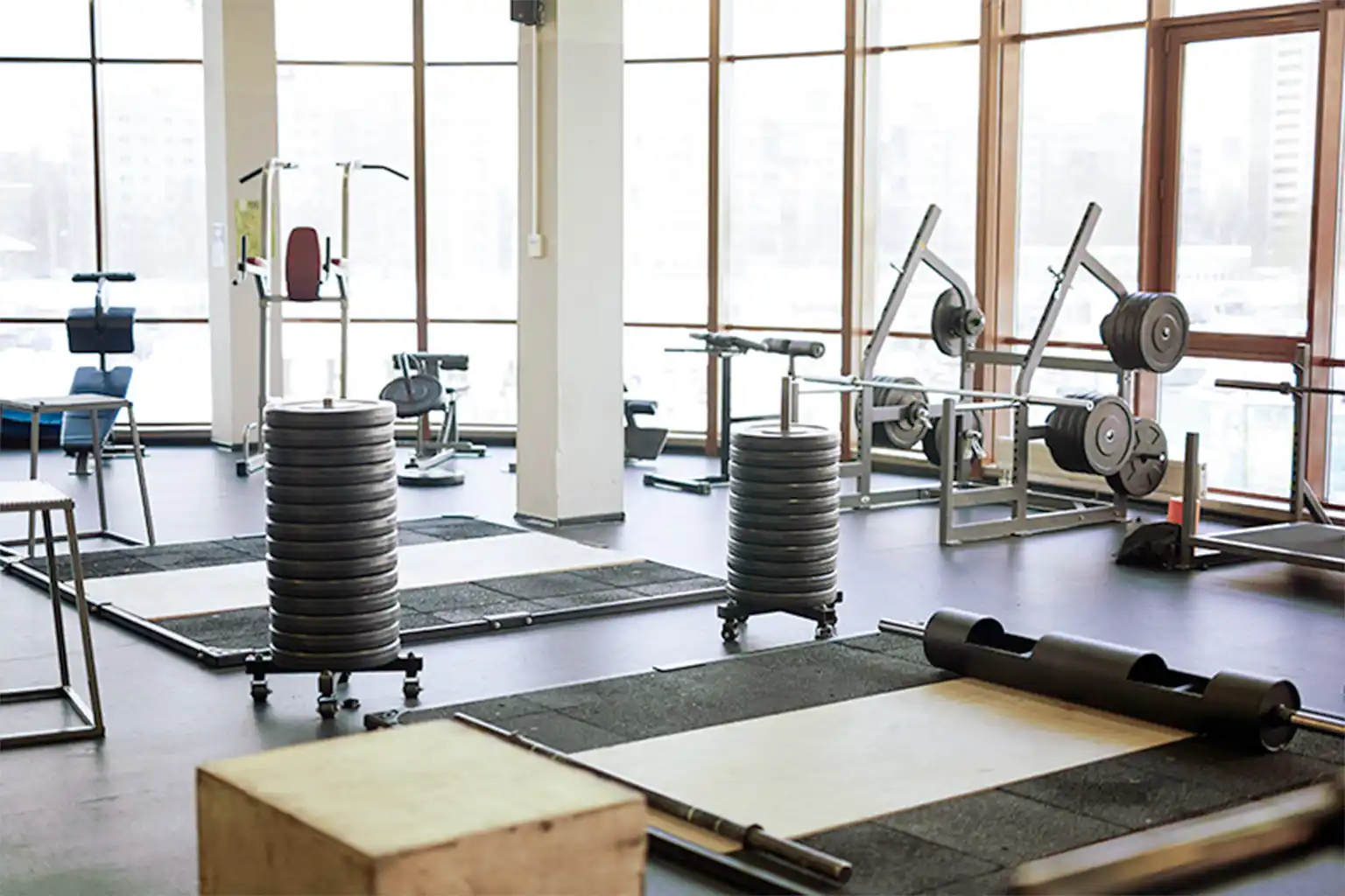Fitnessstudio mit Gewichtsscheiben, Hantelbänken und Trainingsgeräten vor großen Fenstern.