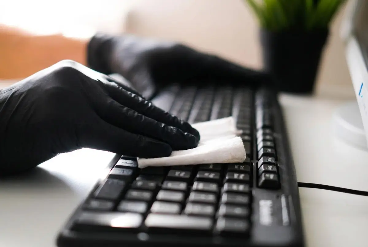 Person wearing black gloves cleaning a computer keyboard with a white cloth.