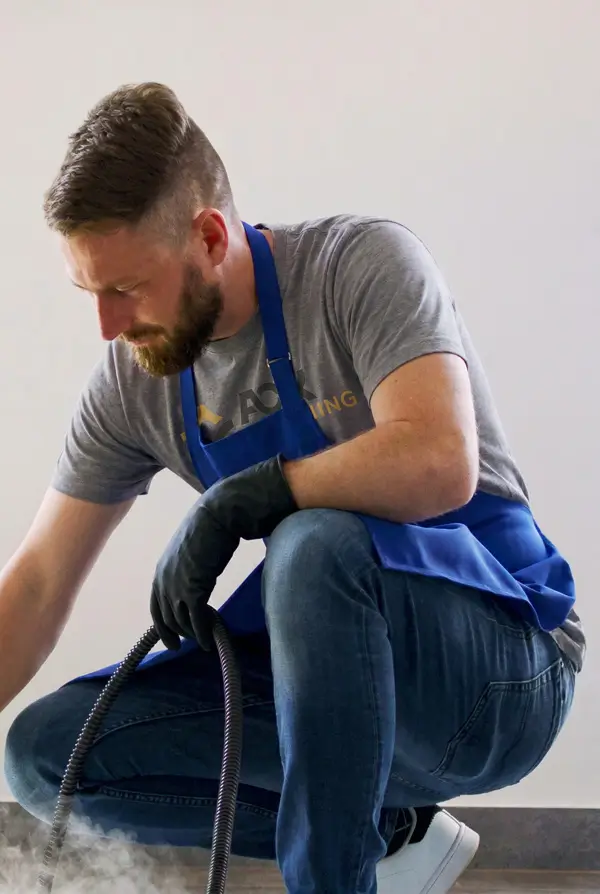Man wearing blue apron and black gloves working with hose nozzle releasing steam on floor.