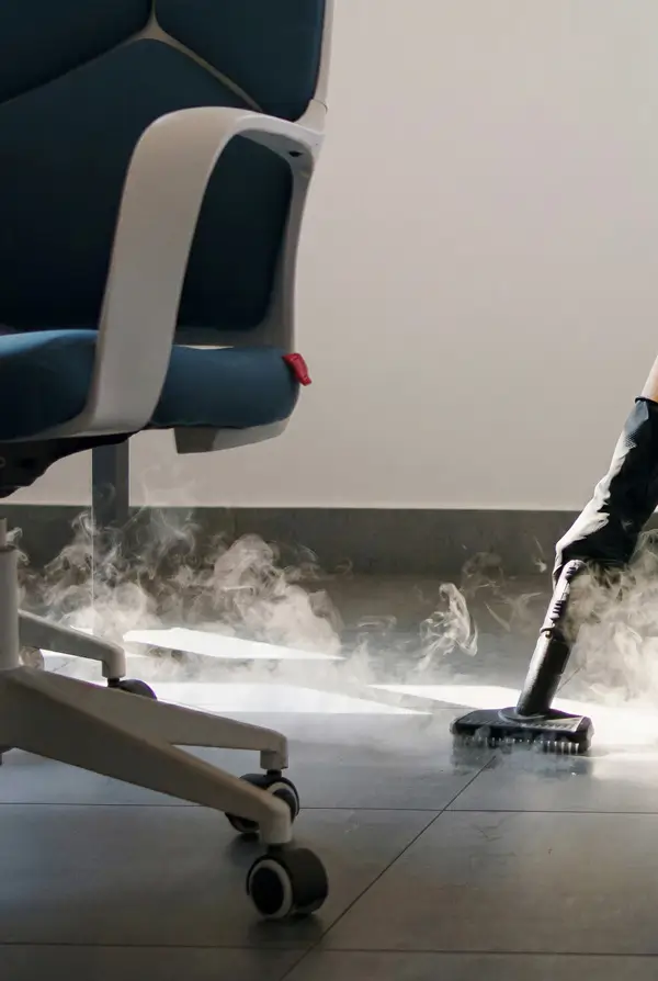 Person using a steam cleaner on dark tiled floor next to an office chair with a dark seat.