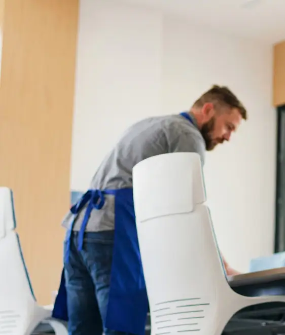 Man in gray shirt and blue apron leaning over a table in a modern office with white chairs.