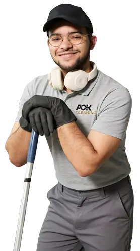 Smiling male cleaner wearing gloves, cap, glasses, headphones around neck, and holding mop handle.