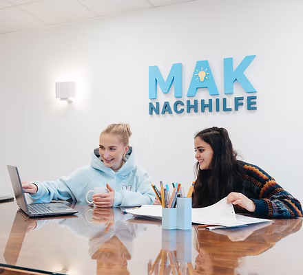 Zwei junge Frauen arbeiten zusammen an einem Laptop am Tisch, MAK Nachhilfe-Logo an der Wand.