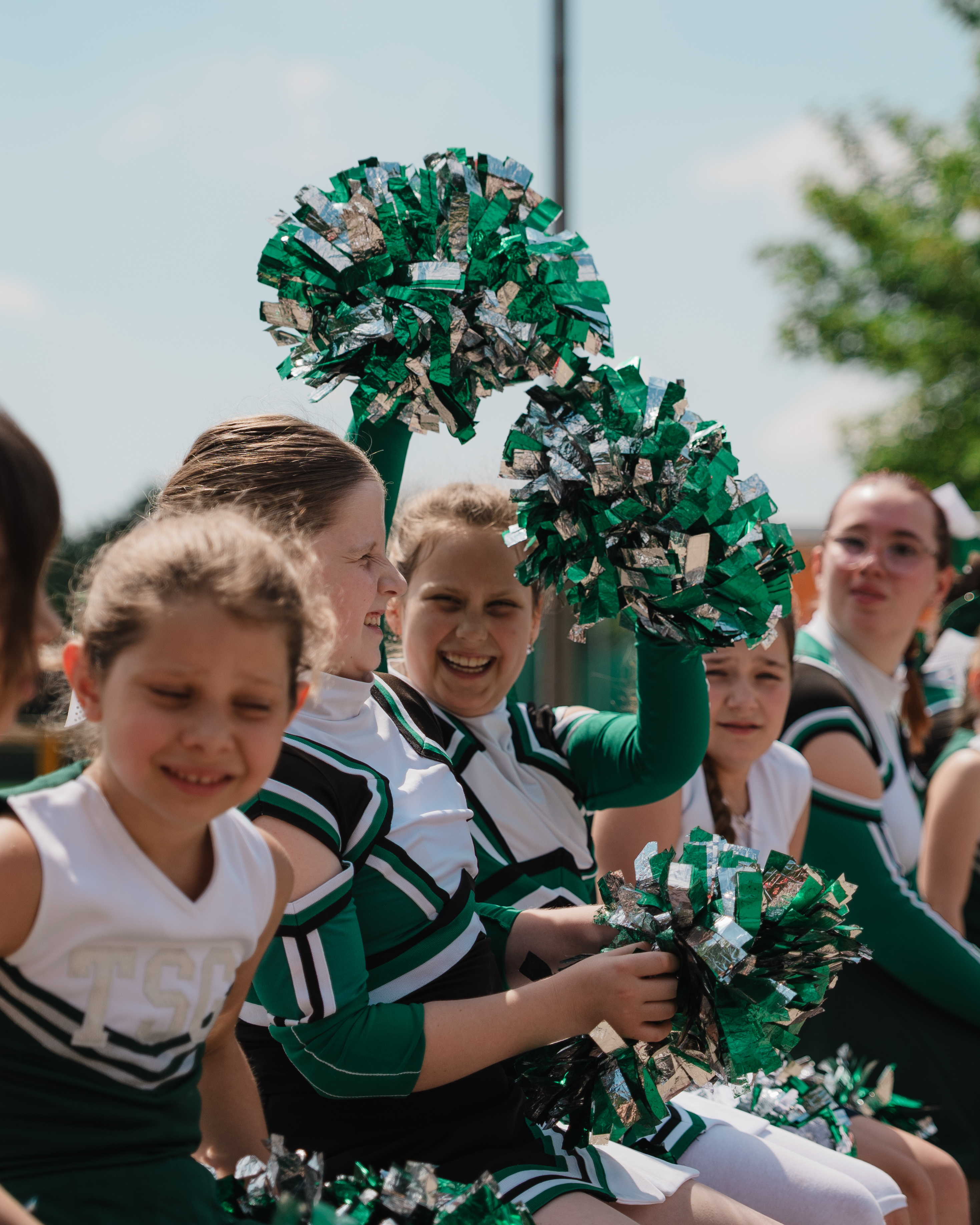 Lachende Cheerleader mit grünen und silbernen Pompons im Freien bei sonnigem Wetter.