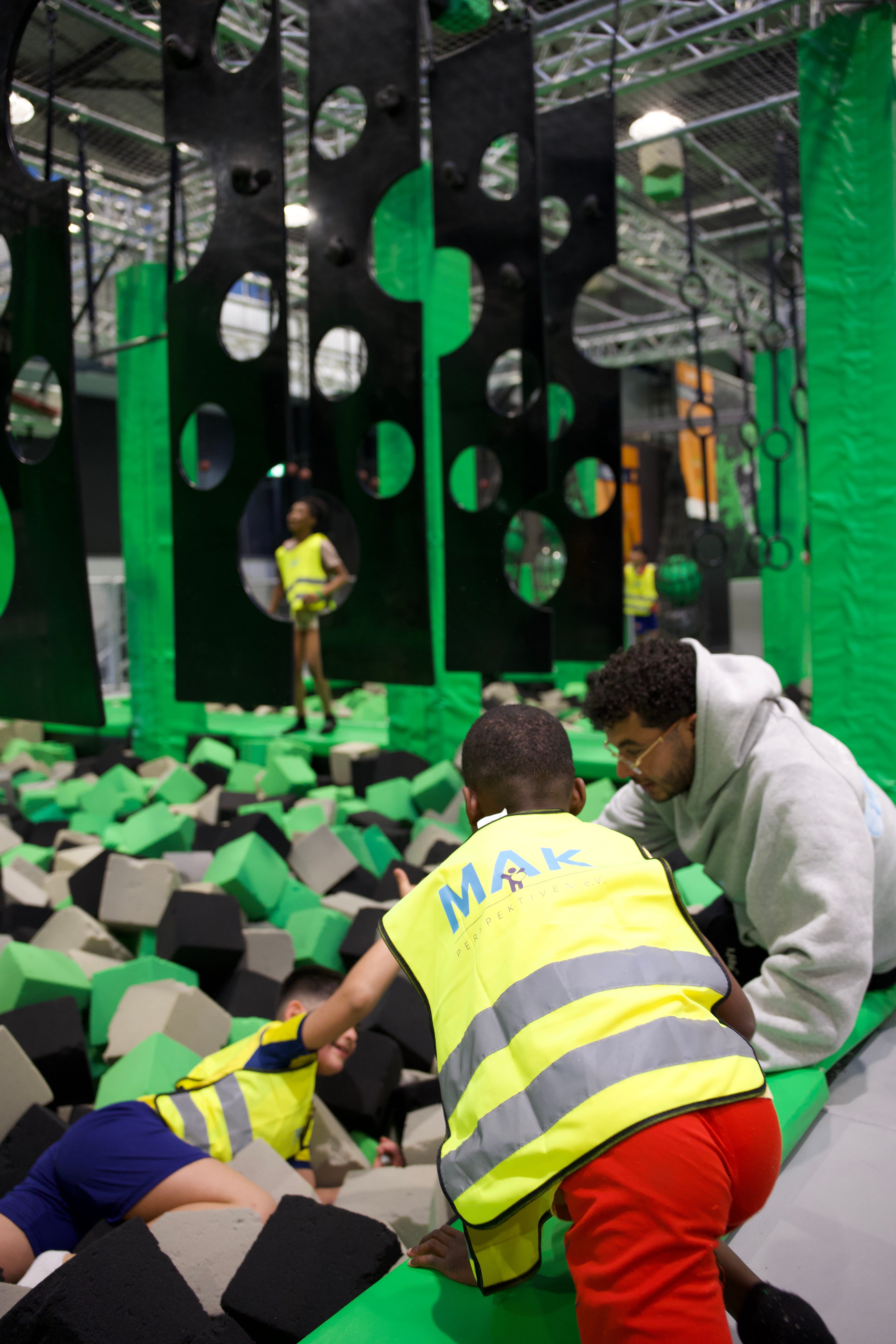 Kinder mit gelben Warnwesten spielen im Schaumstoffbecken eines Kletterparks, ein Erwachsener hilft.