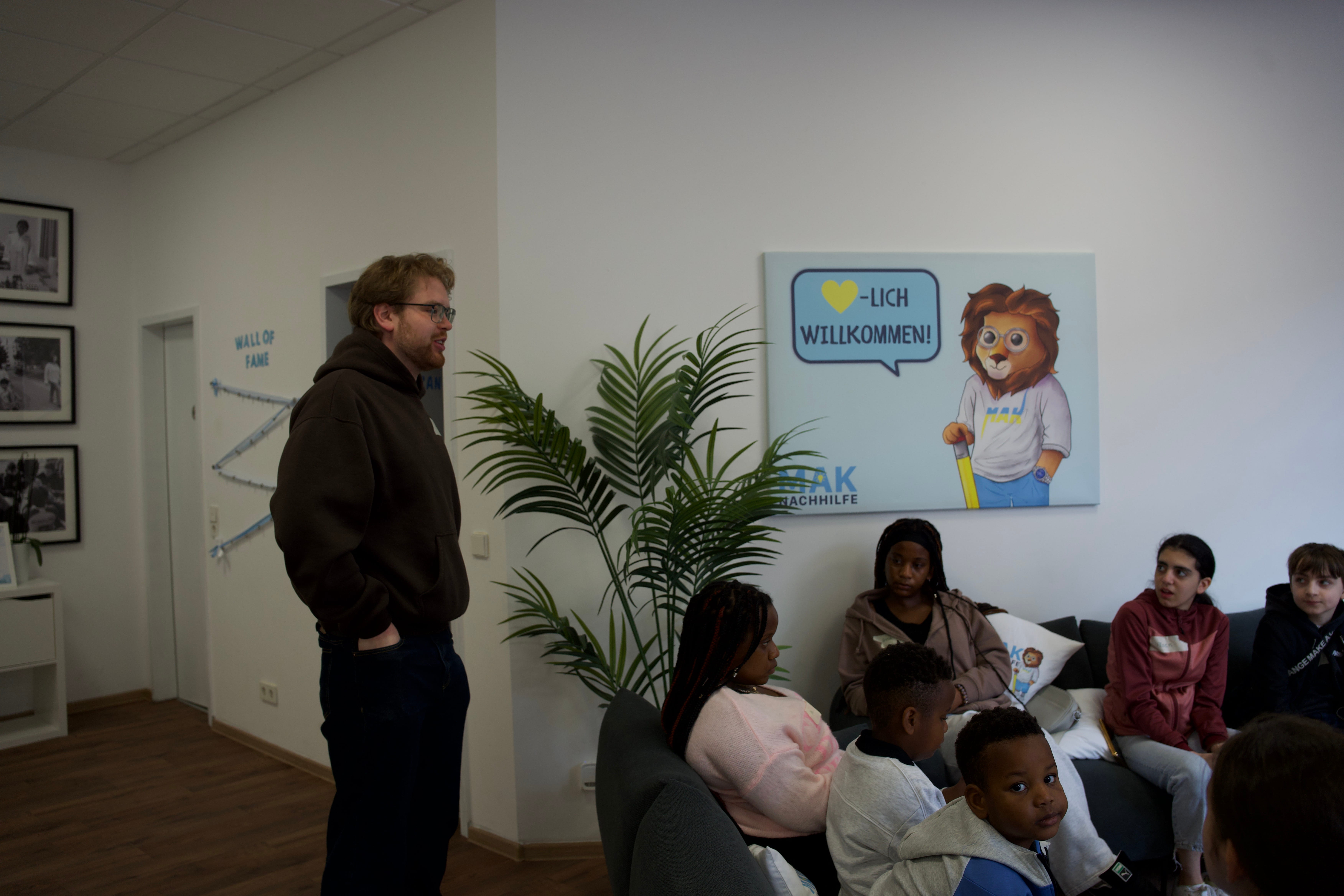 Mann steht vor einer Gruppe Kinder, die auf Sofas in einem Raum mit Willkommensschild sitzen.