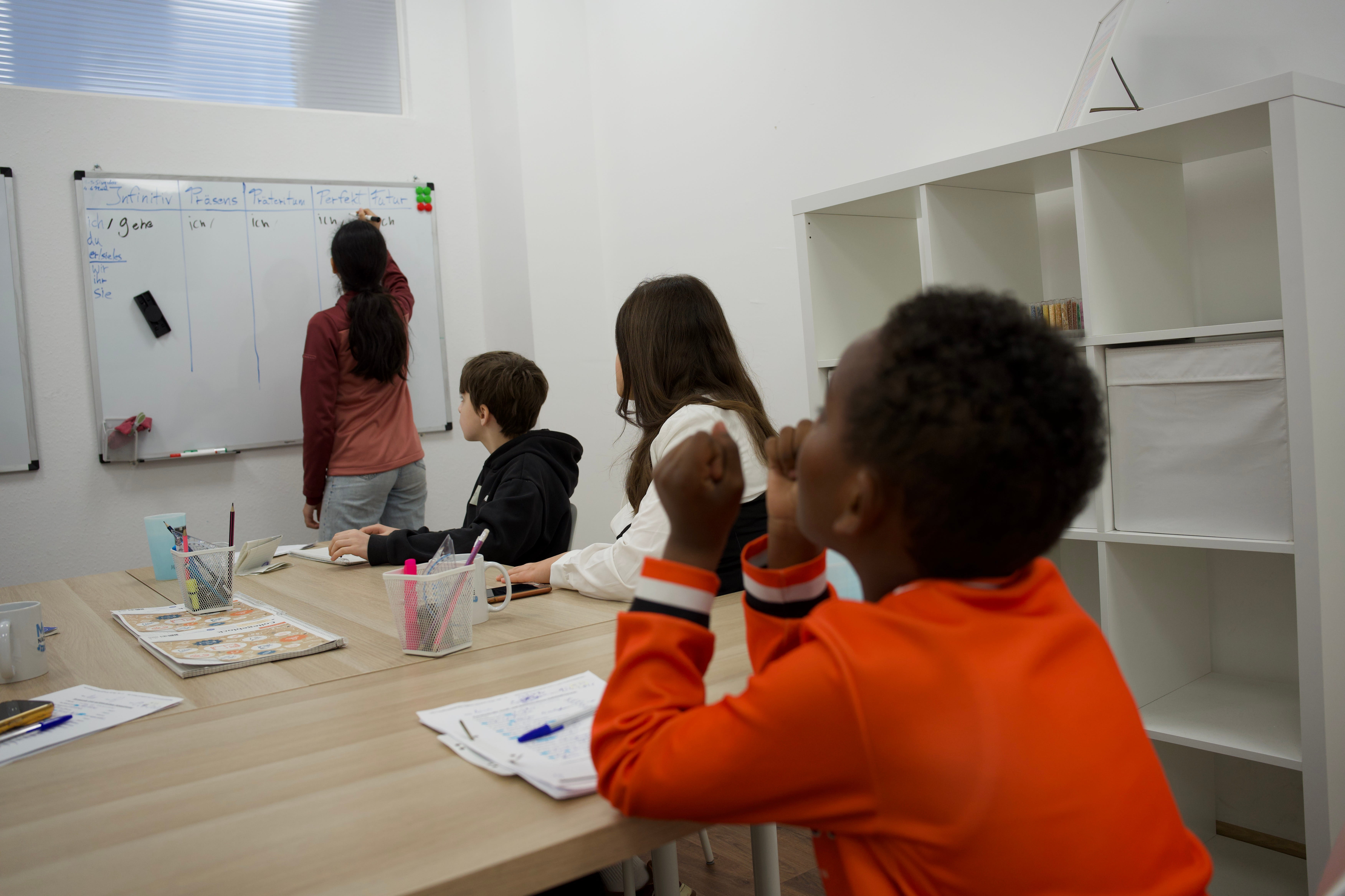 Kinder lernen zusammen in einem Klassenzimmer mit einer Tafel und Bücherregal im Hintergrund.