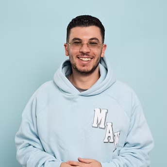 Junger Mann mit Brille und hellblauem Hoodie lächelt vor hellem Hintergrund.
