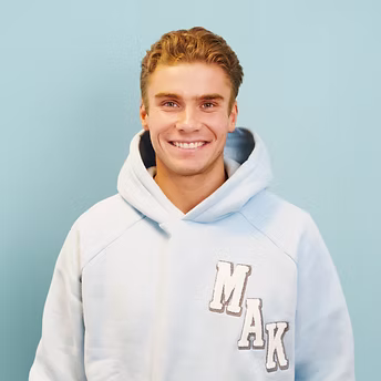 Junger Mann mit hellem Kapuzenpullover lächelnd vor blauem Hintergrund.