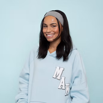 Junge Frau mit langem Haar und grauem Stirnband lächelt in blauem MAK-Hoodie vor hellblauem Hintergrund.