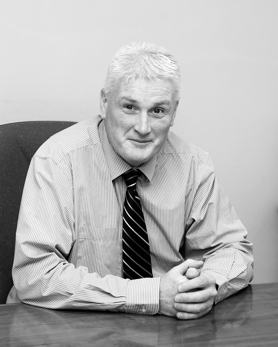 Black and white photo of a man with short white hair seated at a desk, wearing a striped dress shirt and a striped tie, with his hands clasped and resting on the desk, looking directly at the camera and smiling slightly.