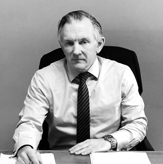Black and white photo of a middle-aged man with slicked-back hair wearing a light-colored dress shirt and striped tie, seated at a desk with papers, looking directly at the camera.