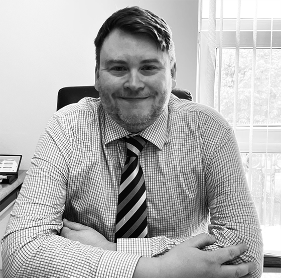 Smiling man with short hair and beard, wearing a checkered shirt and striped tie, sitting at a desk with arms crossed in front, with a window and blinds in the background.
