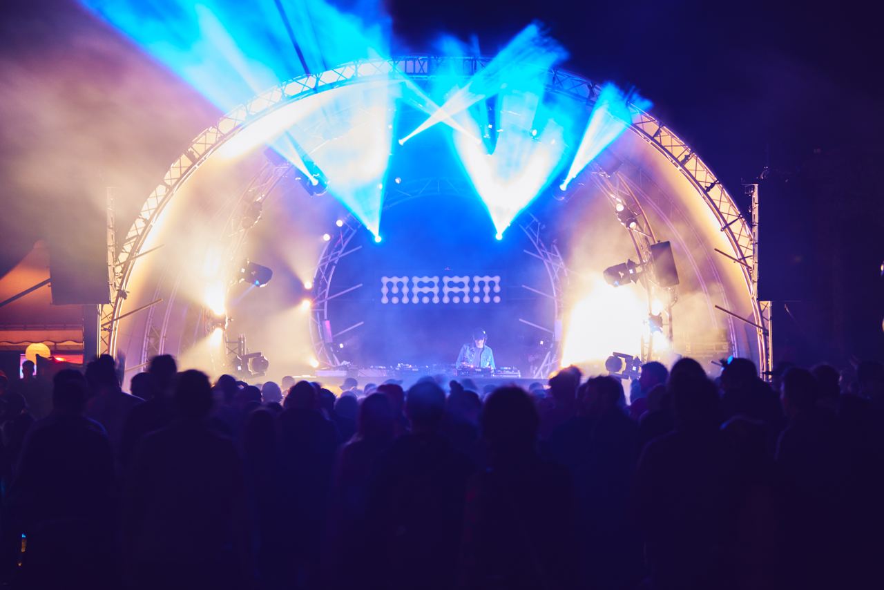DJ che suona su un palco illuminato con uno schermo MAAM e pubblico in silhouette.