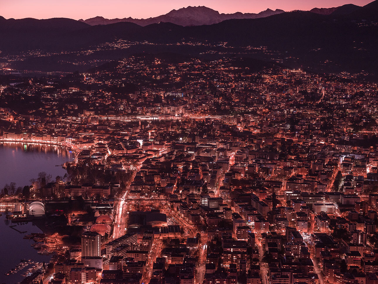 Veduta notturna della città di Lugano illuminata con montagne sullo sfondo al tramonto.