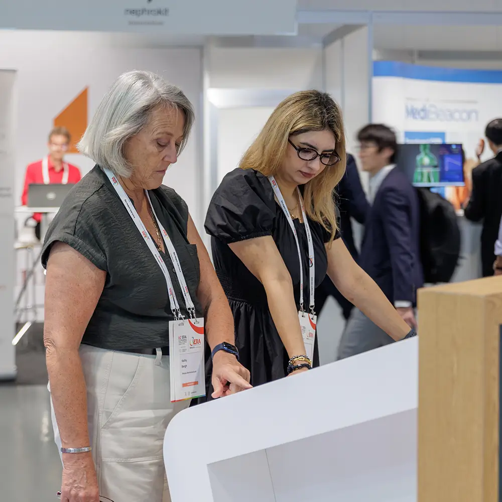 Two women using an interactive touchscreen kiosk at a busy professional event or conference.