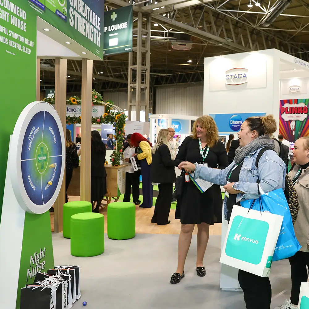People at a trade show playing a wheel game at a Night Nurse booth with green stools nearby.