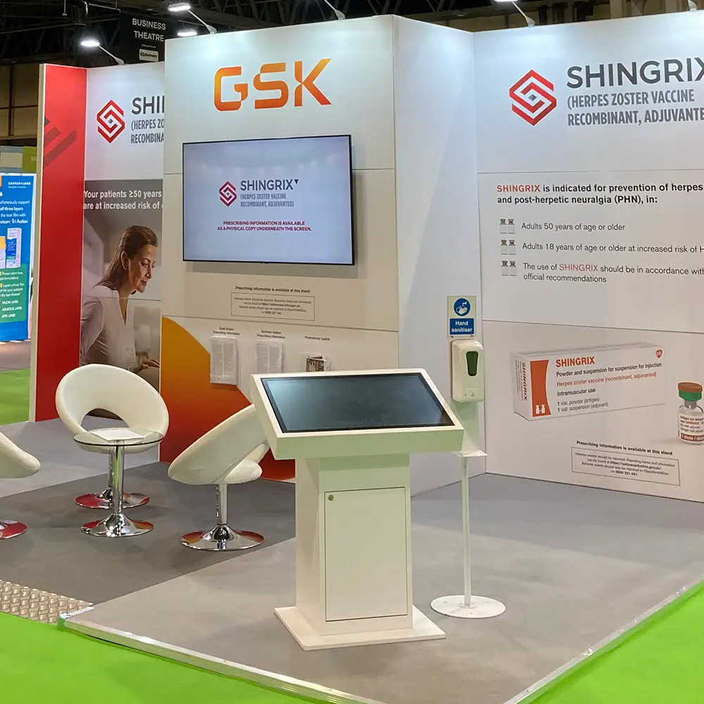 Exhibition booth with GSK and Shingrix vaccine branding, white chairs, touchscreen kiosk, and hand sanitizer.